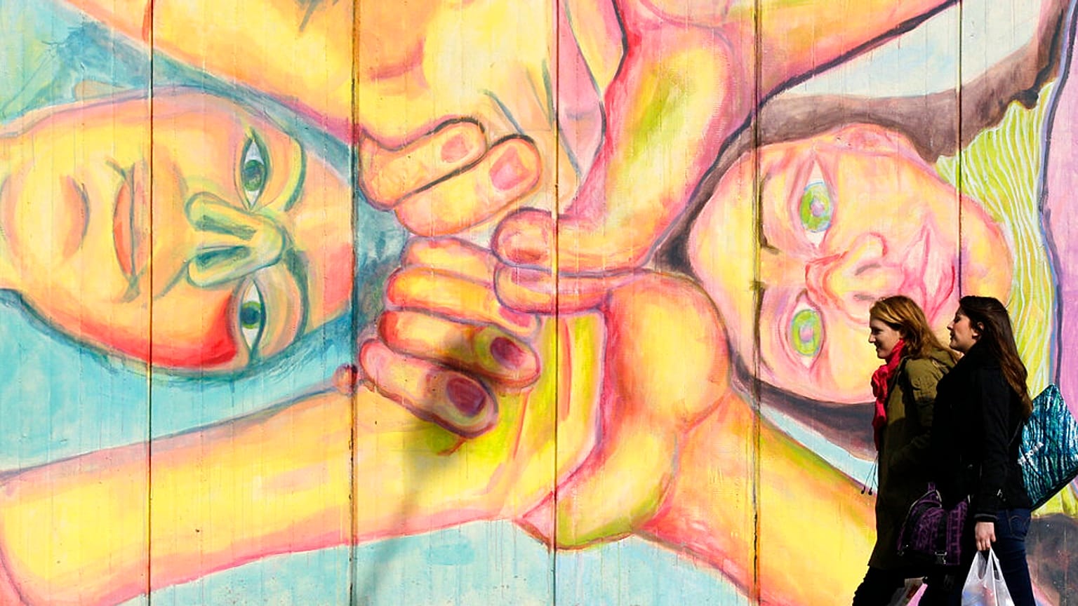 Two women walk past a mural that depicts two women holding hands in Belgrade, Serbia.