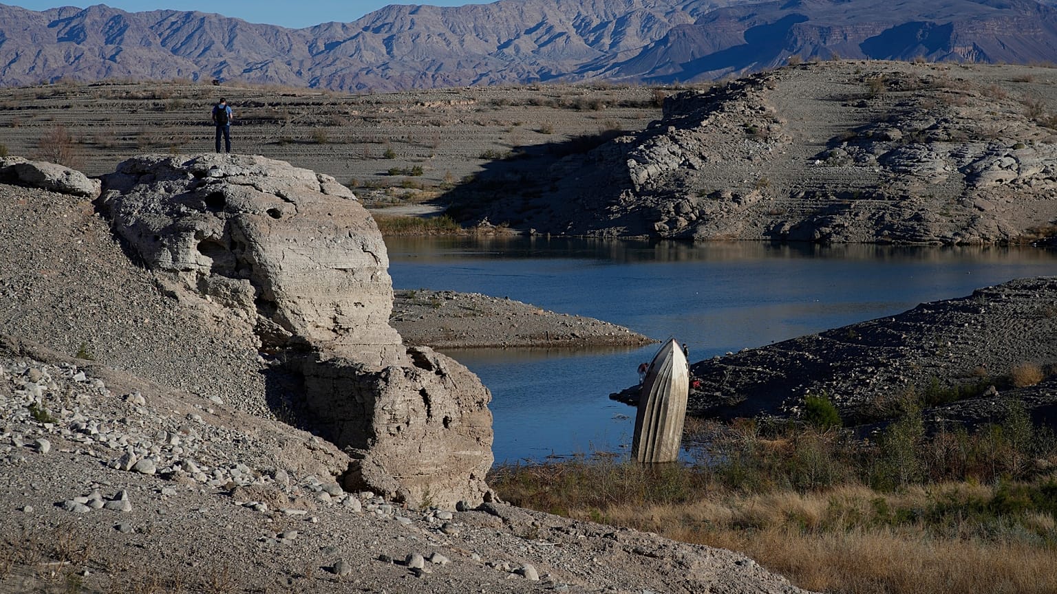 A man stands on a hill overlooking a formerly sunken boat standi.ng upright into the air with its stern buried in the mud along the shoreline of Lake Mead 