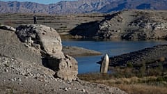 A man stands on a hill overlooking a formerly sunken boat standi.ng upright into the air with its stern buried in the mud along the shoreline of Lake Mead 