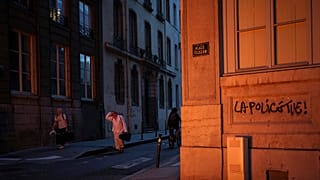 People pass by a wall illuminated by a burning barricade on which is written 'Police kills' in the center of Lyon, central France, Friday, June 30, 2023. 