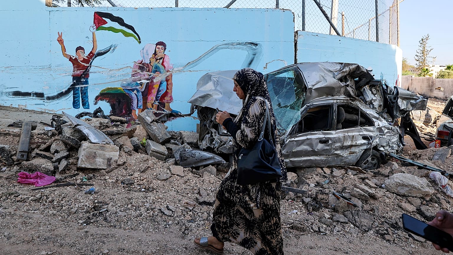 امرأة تمر من طريق مخربة أمام حطام سيارة في إحدى شوارع مدينة جنين في الضفة الغربية المحتلة. 2023/07/05