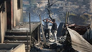 Férfiak egy erdőtűzben kiégett ház előtt az Athéntól északnyugatra fekvő Dervenohoriában 2023. július 19-én.