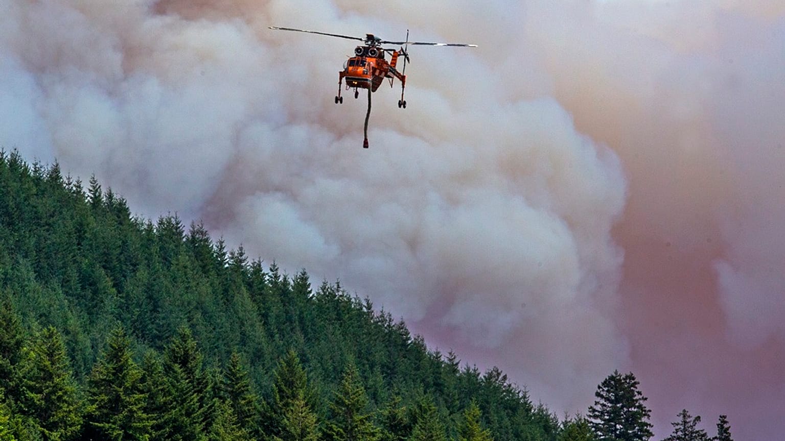 A helicopter flies between the Bedrock Fire and nearby Fall Creek to get a load of water as the wildfire burns east of Eugene, Ore.