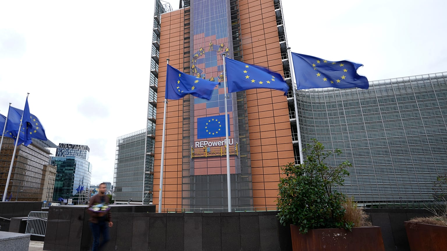 Flags flap in the wind as a man with flowers walks by EU headquarters in Brussels, Wednesday, Sept. 20, 2023.