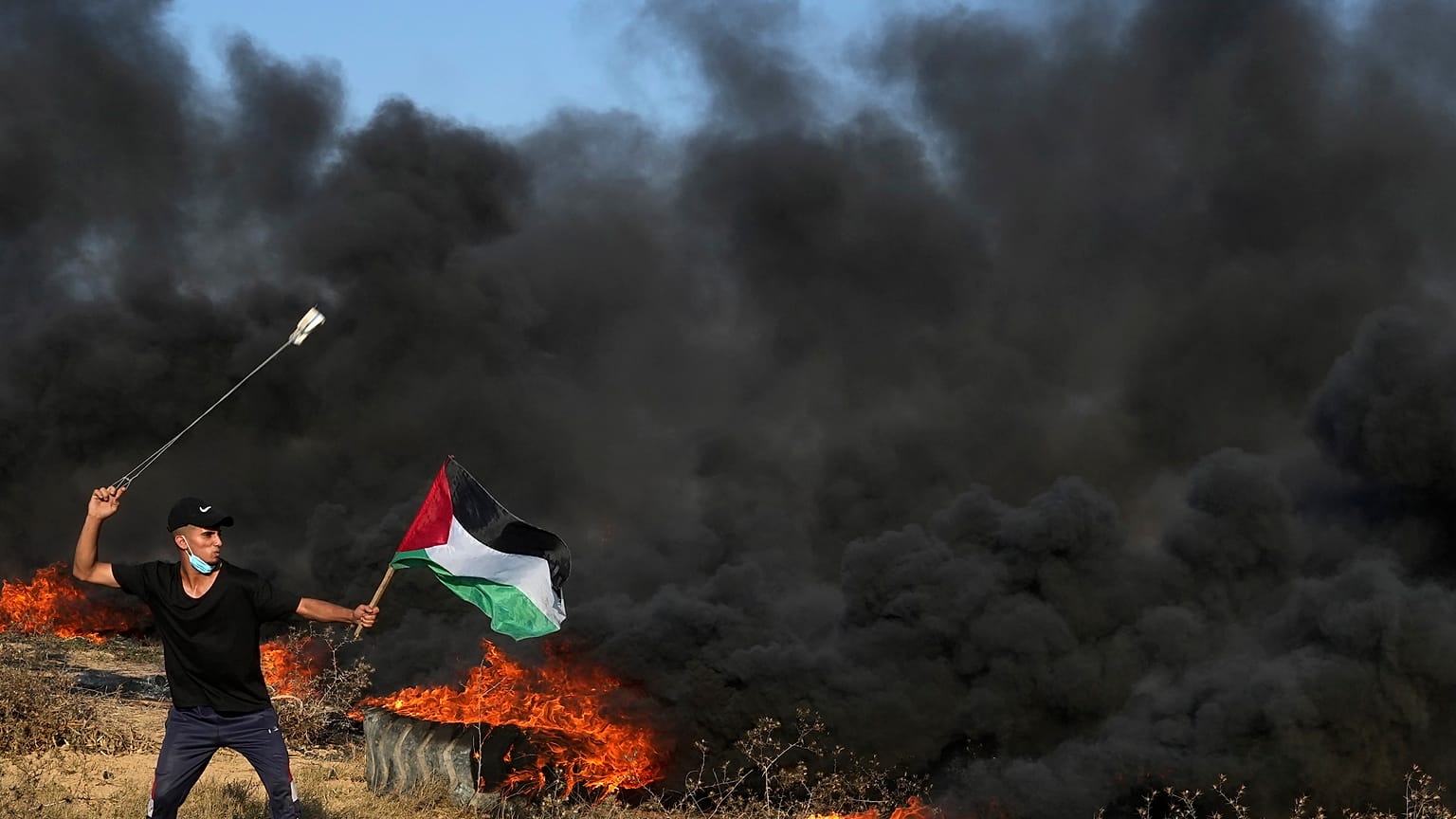 متظاهر فلسطيني يستخدم المقلاع لإلقاء الحجارة بينما يلوح بالعلم الفلسطيني بينما يحرق آخرون الإطارات خلال صدامات مع القوات الإسرائيلية شمال قطاع غزة. 2023/09/22