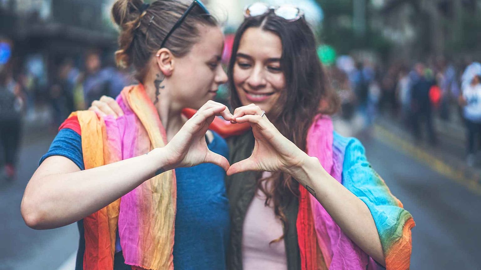 Two women pose with rainbow cloth draped over their shoulders. 