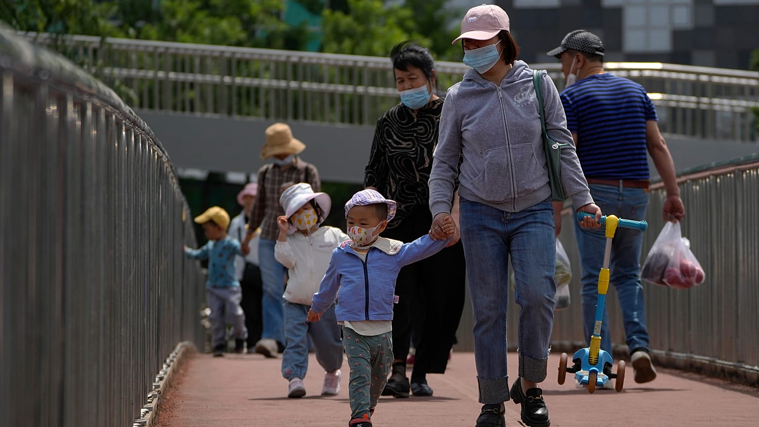 Women wearing face masks walk with masked children as they closing a pedestrian overhead bridge in Beijing, Sunday, May 21, 2023.