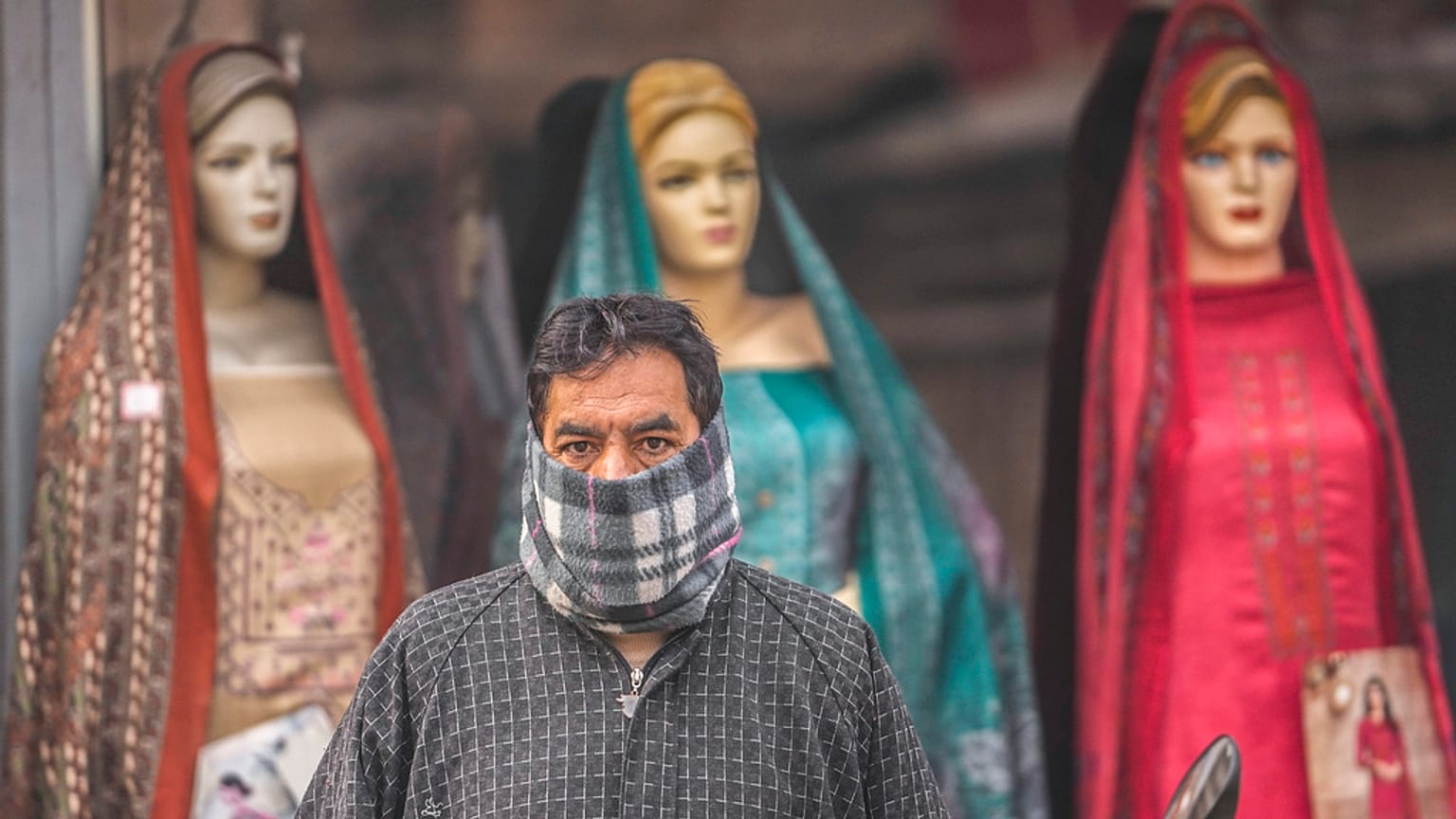 Un uomo ha il volto coperto da un panno di lana mentre attende l'autobus davanti a un negozio di abbigliamento in una giornata fredda a Srinagar