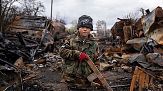 Der kleine Junge Igor, 7, hält ein Spielzeuggewehr aus Holz neben zerstörten russischen Militärfahrzeugen in der Nähe von Tschernihiw, Ukraine.