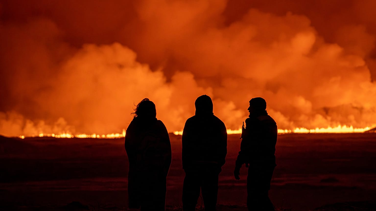 Tres personas observan el cielo iluminado por la erupción de un volcán en Grindavik, en la península islandesa de Reykjanes.