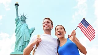 دو بازدیدکننده با پرچم ایالات متحده آمریکا در نزدیکی مجمسه آزادی در نیویورک