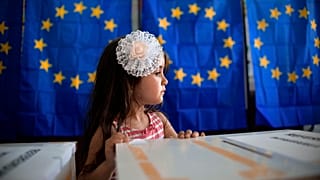 A little girl waits for her mother to vote, backdropped by voting booths with curtains depicting the European Union flag in Baleni, Romania, in 2019.