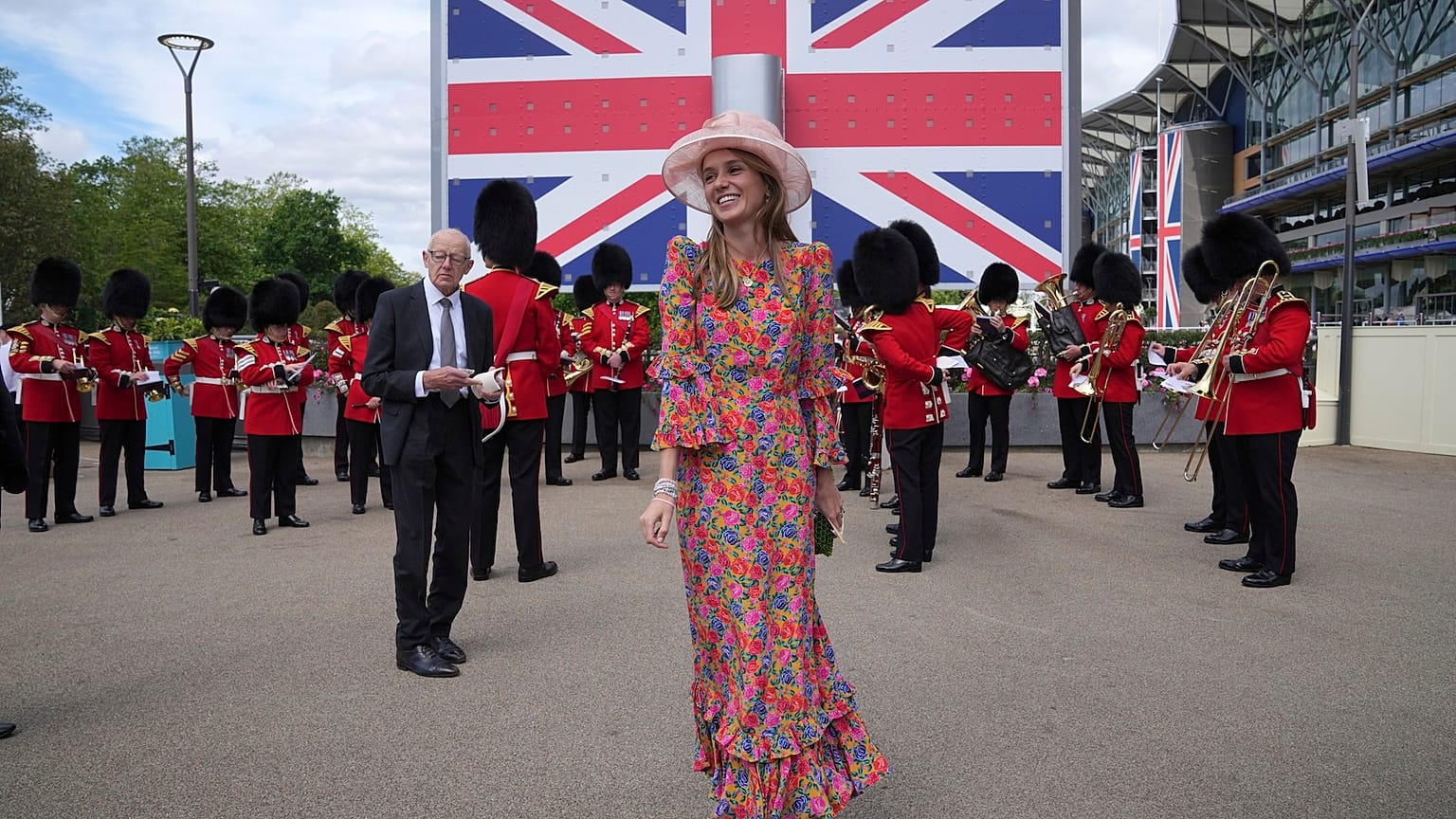 A young lady in hat and floral dress smiles in front of a British flag display as a military ban plays in the background on the second day of Royal Ascot races 
