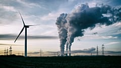 Un aerogenerador, una central eléctrica de carbón con contaminación y torres de alta tensión al fondo.