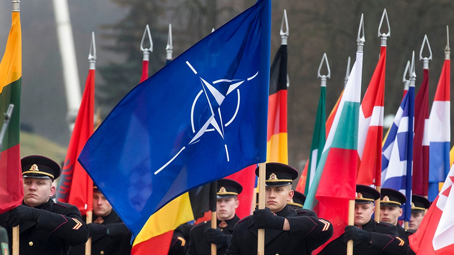 Bandeira da NATO com as bandeiras dos países membros a serem flanqueadas durante uma marcha militar