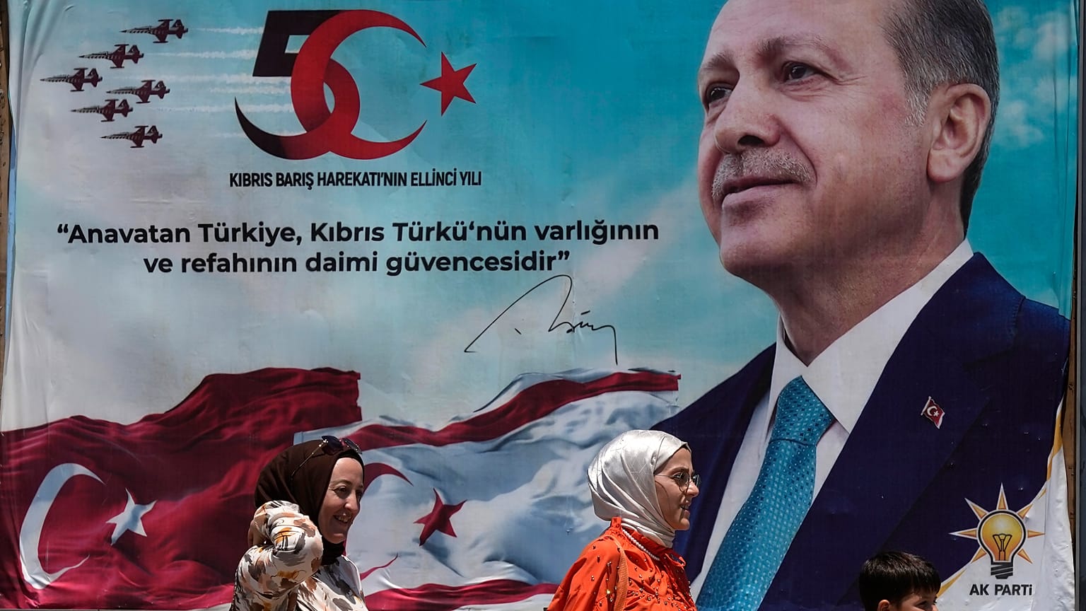 Mulheres turcas e uma criança caminham em frente a um cartaz do Presidente Recep Tayyip Erdoğan após uma parada militar no norte de Chipre, 20 de julho de 2024