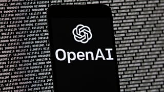 The OpenAI logo appears on a mobile phone in front of a computer screen with random binary data.