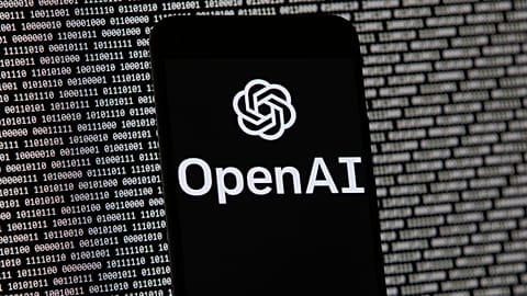 The OpenAI logo appears on a mobile phone in front of a computer screen with random binary data.