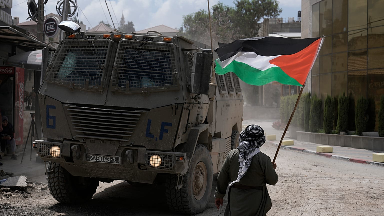 رجل يلوح بعلم فلسطيني بينما تتحرك مركبة مدرعة إسرائيلية في أحد الشوارع خلال عملية عسكرية في مخيم نور شمس للاجئين بالضفة الغربية، طولكرم، الخميس 29 أغسطس/آب