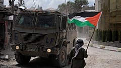 رجل يلوح بعلم فلسطيني بينما تتحرك مركبة مدرعة إسرائيلية في أحد الشوارع خلال عملية عسكرية في مخيم نور شمس للاجئين بالضفة الغربية، طولكرم، الخميس 29 أغسطس/آب