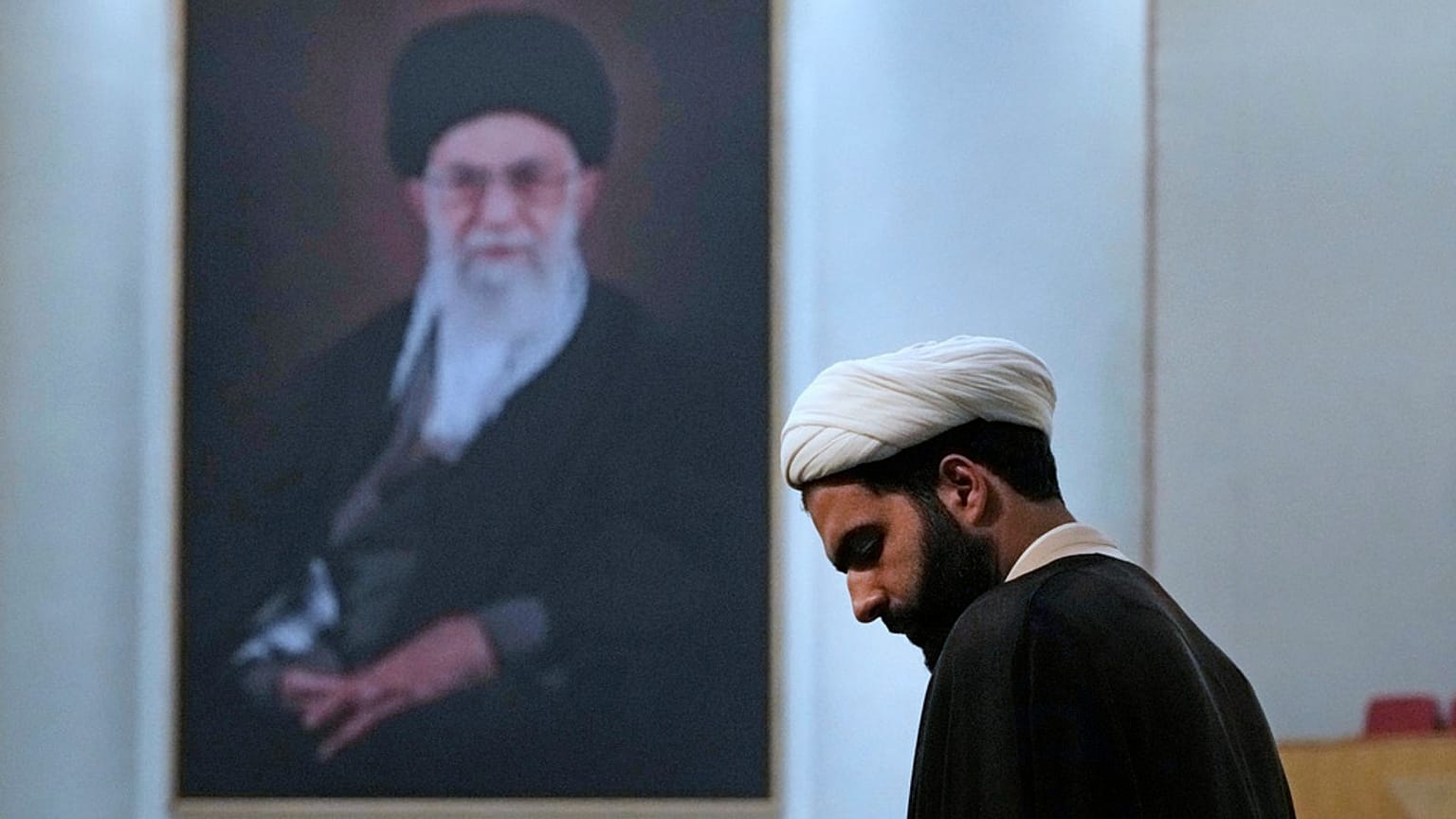 یک روحانی هوادار حکومت ایران در برابر عکس علی خامنه‌ای، رهبر جمهوری اسلامی