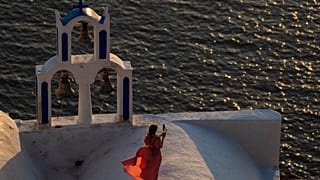 A woman takes pictures as she stands on the top of a Greek Orthodox Church while the sun sets in the Aegean Sea, in Santorini