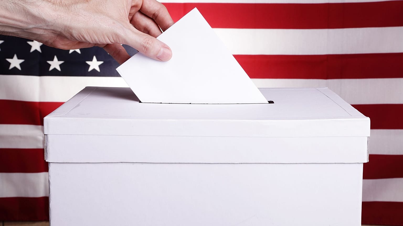 Un hombre deposita su voto con una bandera estadounidense detrás.