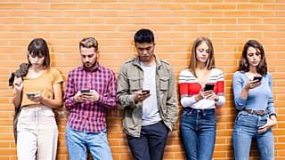 Bir tuğla duvara yaslanmış, telefonlarıyla meşgul bir dizi genç insan. 
