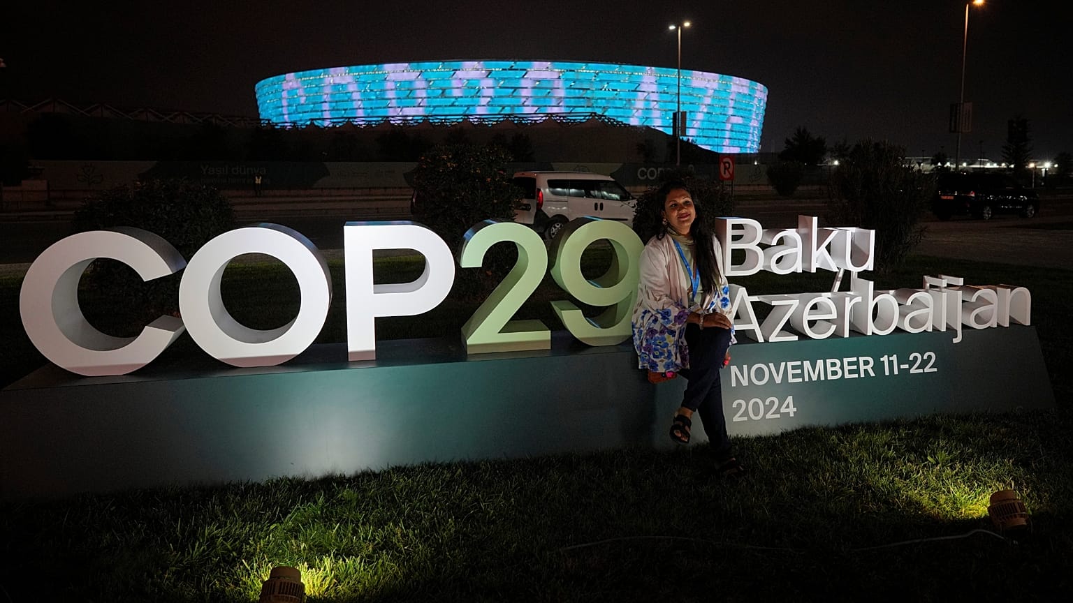 A woman poses for a photo with the Baku Olympic Stadium in the background as it is illuminated at COP29.