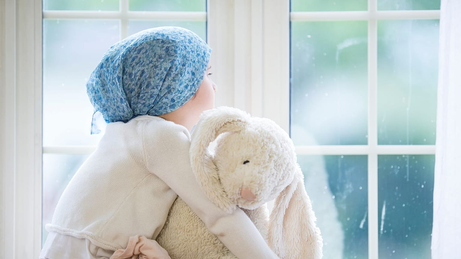 Una niña con cáncer mira a través de la ventana abrazando a su oso.