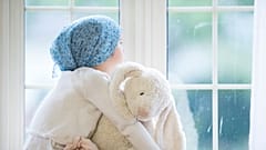 Una niña con cáncer mira a través de la ventana abrazando a su oso.