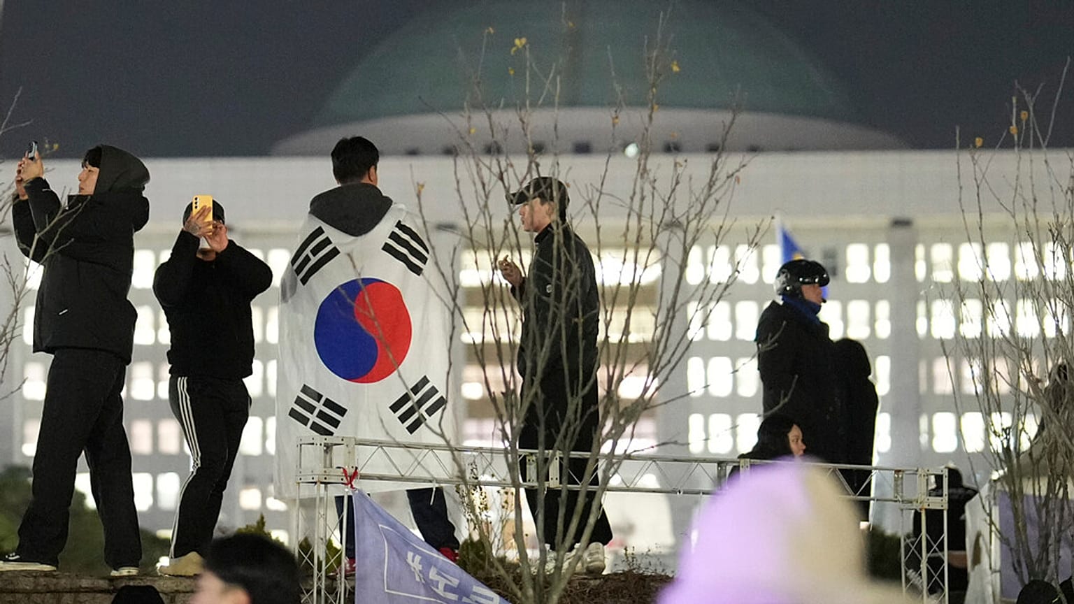 Un uomo che indossa una bandiera nazionale si trova sul muro dell'Assemblea nazionale a Seul, Corea del Sud, mercoledì 4 dicembre 2024.