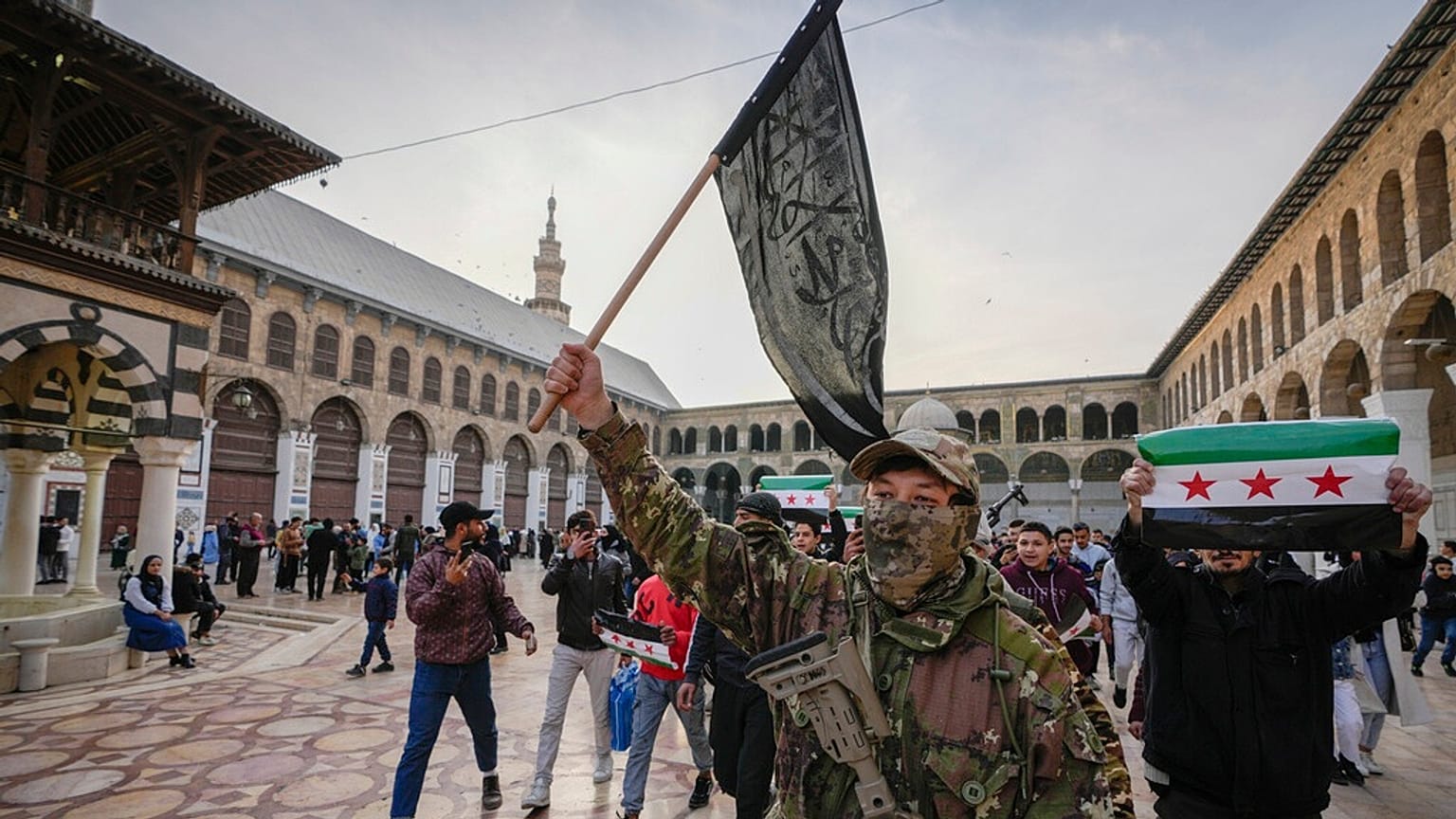 Maskeli bir muhalif savaşçı, Suriye'nin surlarla çevrili eski şehri Şam'daki Emevi Camii'nin avlusunda Heyet Tahrir el Şam'ın (HTŞ) bayrağını taşıyor, 2024.