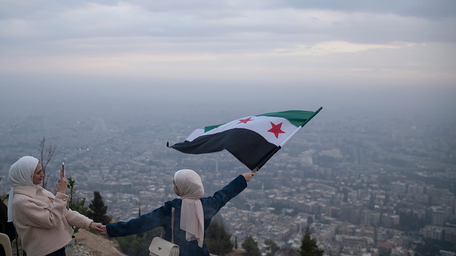 امرأة شابة تقف لالتقاط صورة وهي تلوح بعلم "الثورة" السورية في قمة على جبل قاسيون في دمشق، سوريا، السبت 21 ديسمبر 2024.