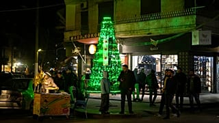 سوريون يقفون بجانب شجرة عيد الميلاد المضاءة المزينة بالعلم السوري ”الثوري“ في مدينة حلب، سوريا، السبت، 14 ديسمبر/كانون الأول 2024. 