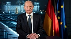Kanclerz Niemiec Olaf Scholz siedzi na tle Reichstagu oświetlonego reflektorami podczas sesji zdjęciowej po nagraniu przemówienia noworocznego.