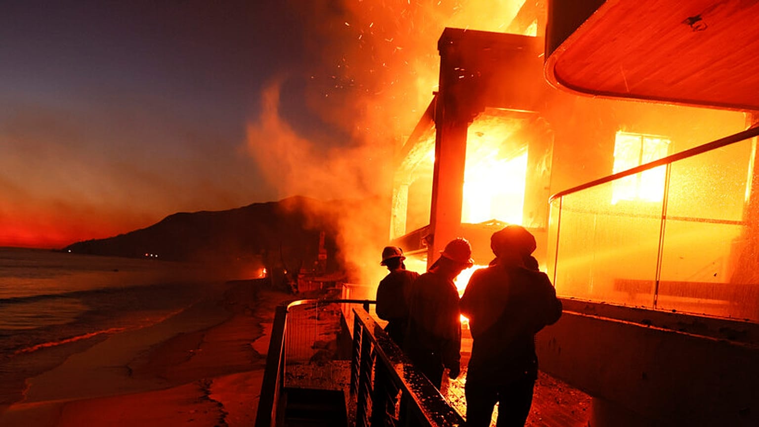 Os bombeiros trabalham a partir de um convés enquanto o incêndio em Palisades queima uma propriedade em frente à praia na quarta-feira, em Malibu, Califórnia