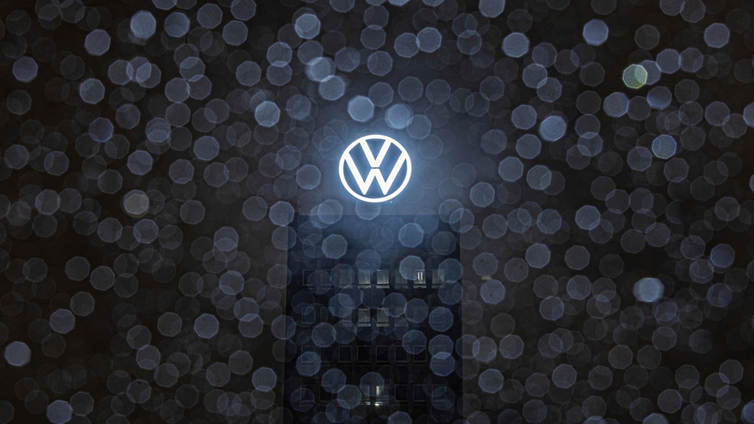 Yağmur damlaları bir otomobilin ön camında toplanırken Volkswagen marka kulenin çatısındaki logo arkada parlıyor. Wolfsburg, Almanya. 9 Aralık 2024.