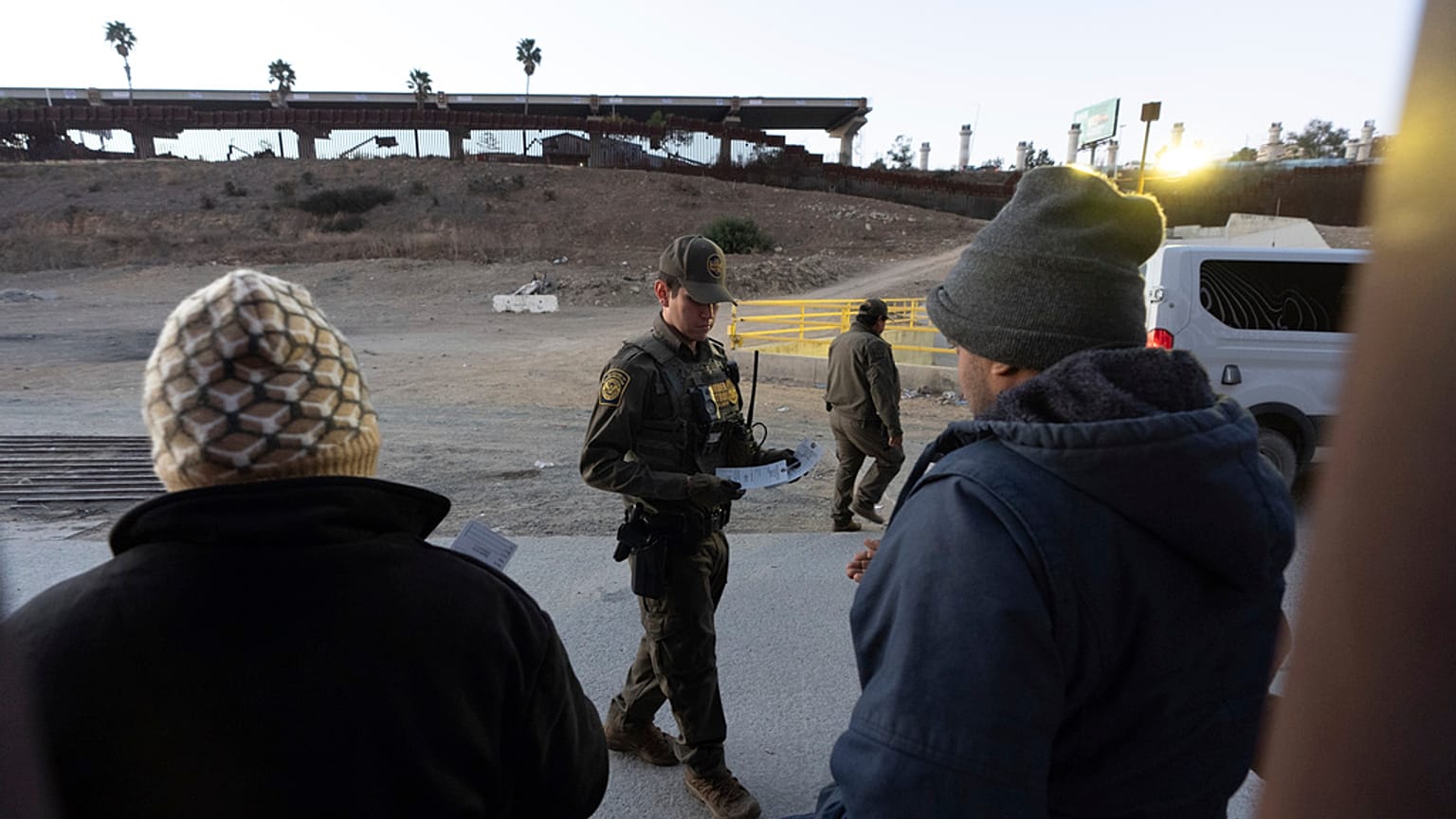 Un agente de la Patrulla Fronteriza reparte etiquetas de equipaje a un grupo de migrantes que quieren solicitar asilo entre dos muros fronterizos que separan a México y EE.UU.