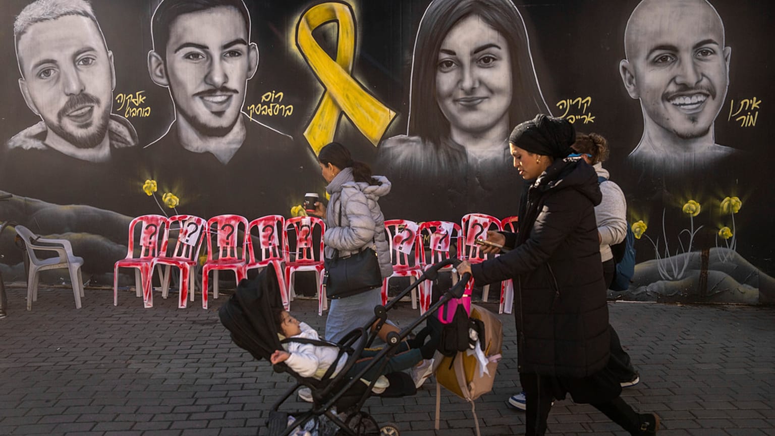 İnsanlar 24 Ocak 2025 Cuma günü Kudüs'te, Gazze Şeridi'nde Hamas tarafından tutulan rehinelerin portrelerinden oluşan bir duvar resmi ve enstalasyonun önünden yürüyerek ve koşarak geçiyor. 