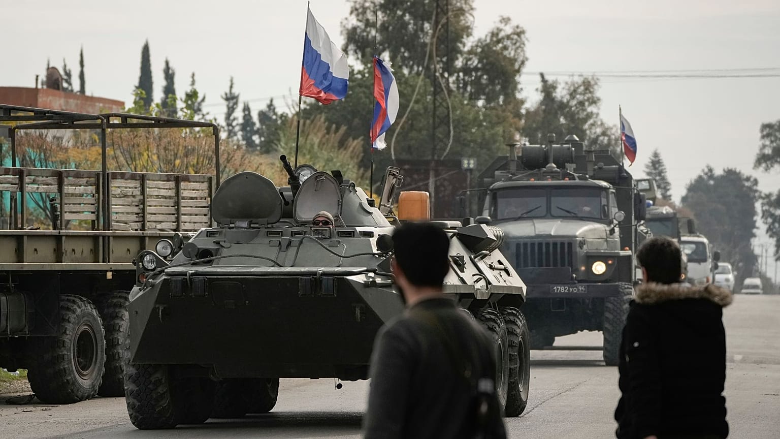 FILE - Combattenti siriani osservano i veicoli blindati russi che passano vicino alla base aerea di Hmeimim, una base aerea siriana attualmente gestita dalla Russia, 16 dicembre 2024. 