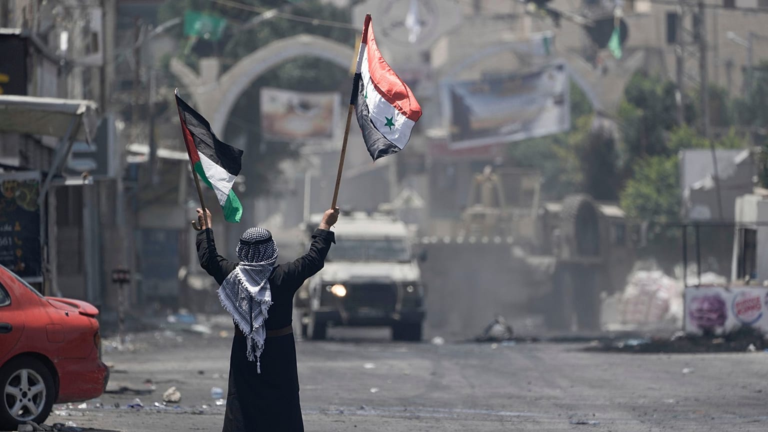 یک مرد فلسطینی با پرچم‌های فلسطین و سوریه در مقابل خودروی ارتش اسرائیل در حمله نظامی به اردوگاه آوارگان جنین در کرانه باختری اشغالی به تاریخ چهارم ژوئیه ۲۰۲۳