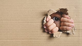 Imagen de un niño que se esconde y observa, mientras permanece escondido detrás de un trozo de cartón.