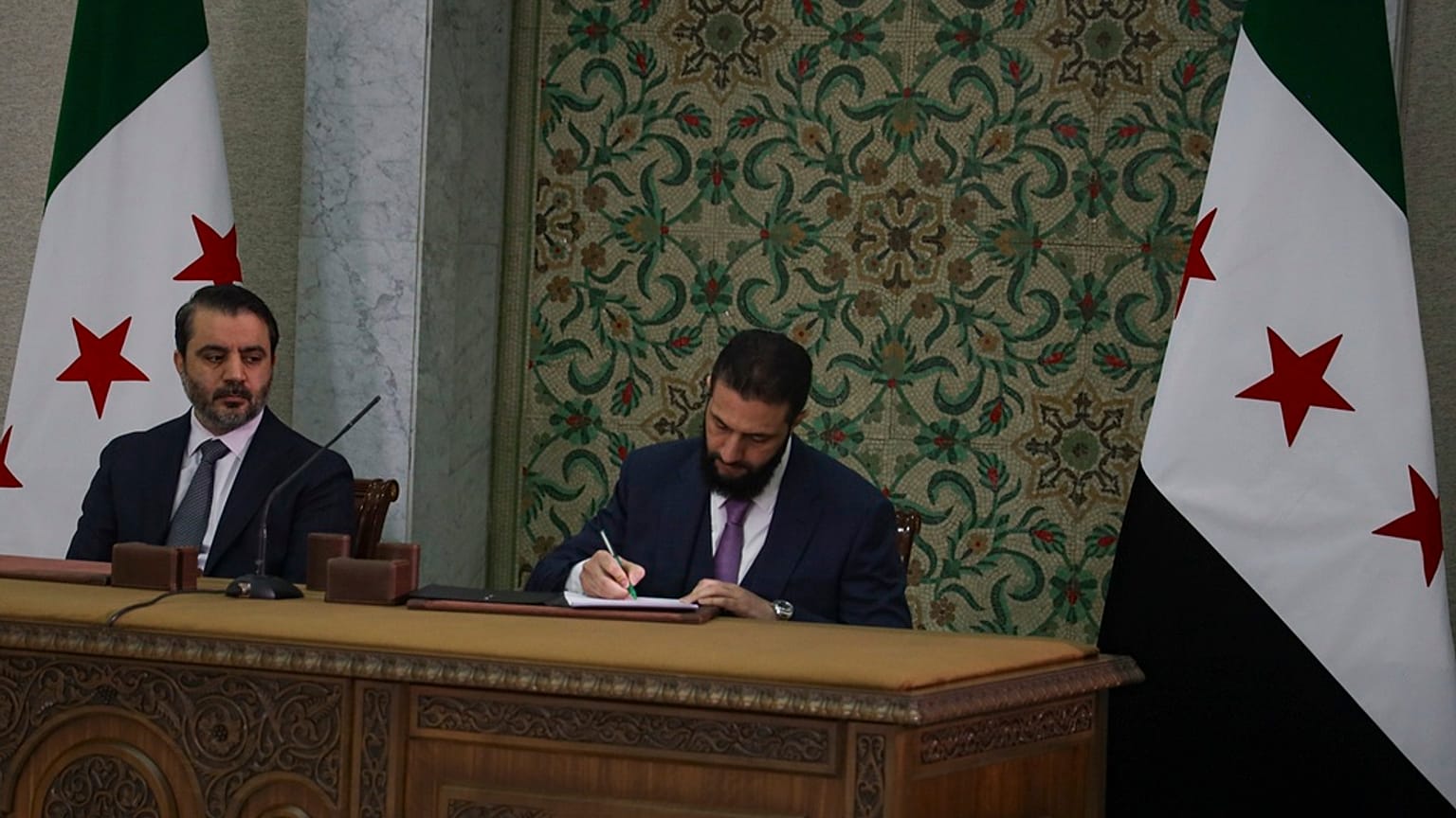 الرئيس السوري المؤقت أحمد الشرع، في الوسط، يوقع على دستور مؤقت للبلاد في دمشق، سوريا، الخميس 13 مارس/آذار 2025. إلى اليسار وزير الخارجية أسعد حسن الشيبان