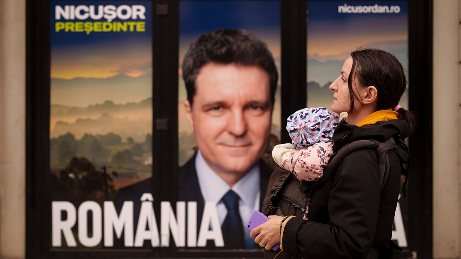 Kucağında bebek taşıyan bir kadın, Bükreş Belediye Başkanı ve Mayıs ayında yapılacak seçimlerde cumhurbaşkanı adayı olan Nicusor Dan'ın posterinin yanında yürürken, Bükreş, 25 Mart 2025
