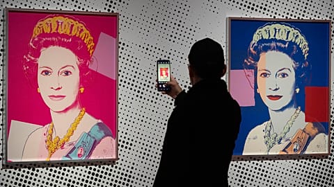 A man takes an image of a screen print depicting Queen Elizabeth II, one in a series of sixteen prints of four queens titled Reigning Queens, 1985, by Andy Warhol