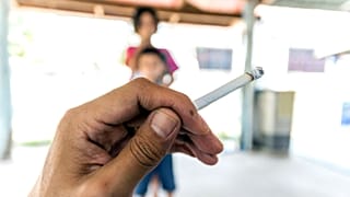 Una mano sostiene un cigarrillo con niños al fondo.
