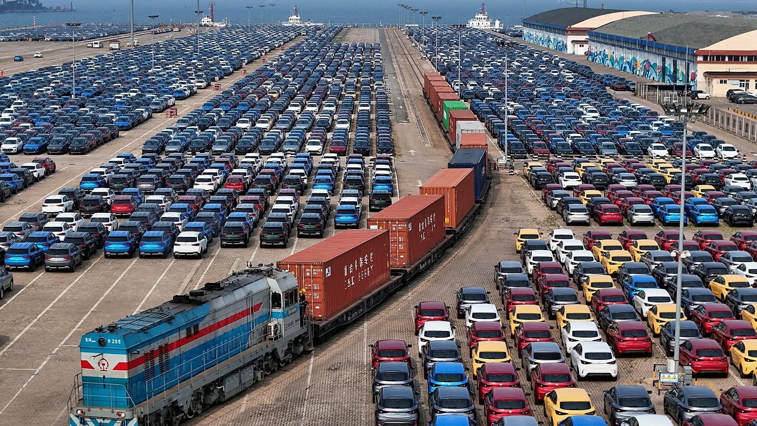 A cargo train moves past new cars waiting for shipment in a port, in Yantai in east China's Shandong province, 7 May 2025