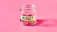 Um frasco de vidro com algumas moedas e uma etiqueta com a inscrição "Budget" sobre um fundo cor-de-rosa.
