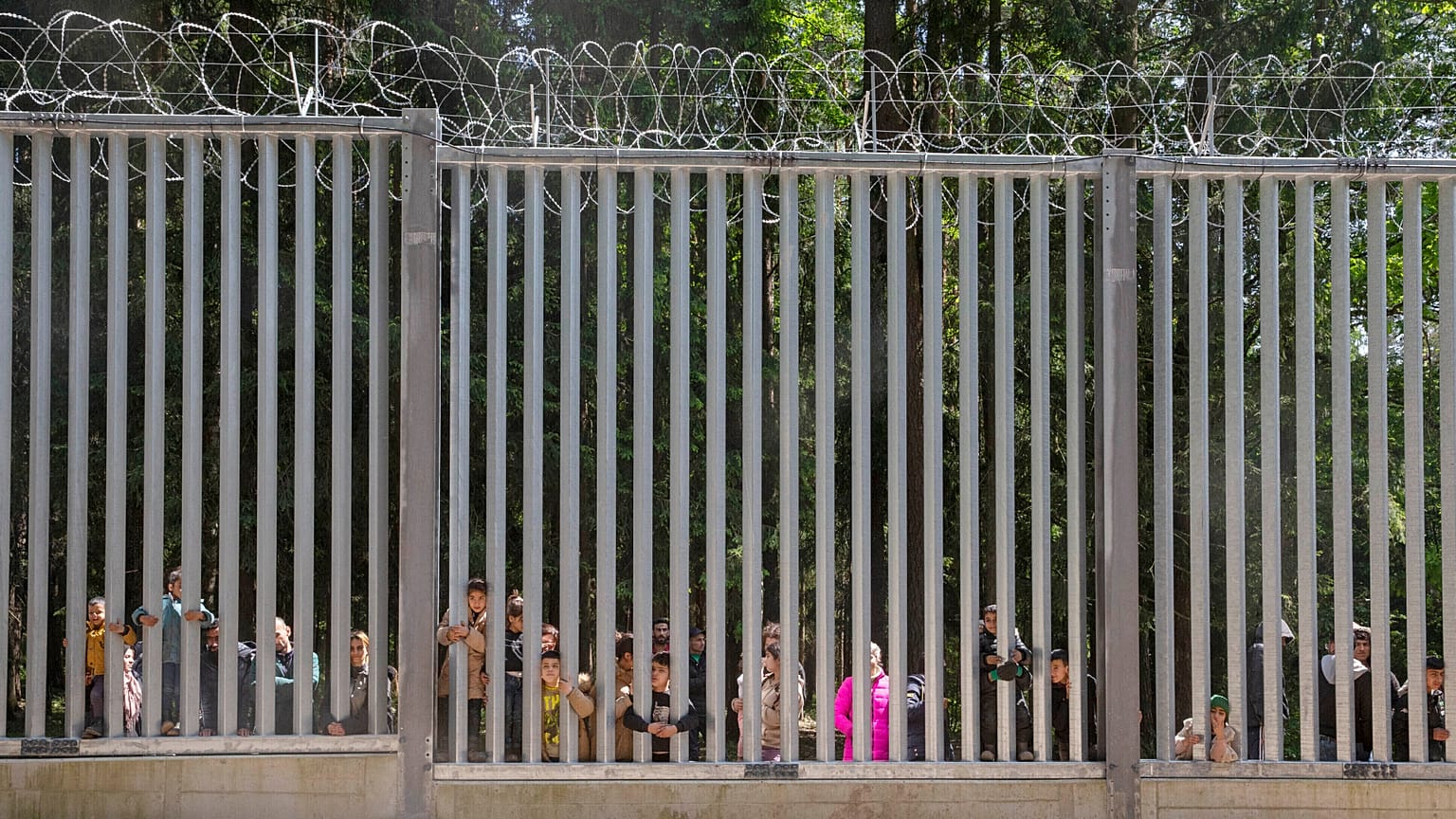 A lengyelországi Bialowiezában 2023. május 28-án, vasárnap egy mintegy 30 fős, menedéket kérő migránscsoport tagjai a Lengyelország által a határon épített fal korlátján keresztül néznek át.